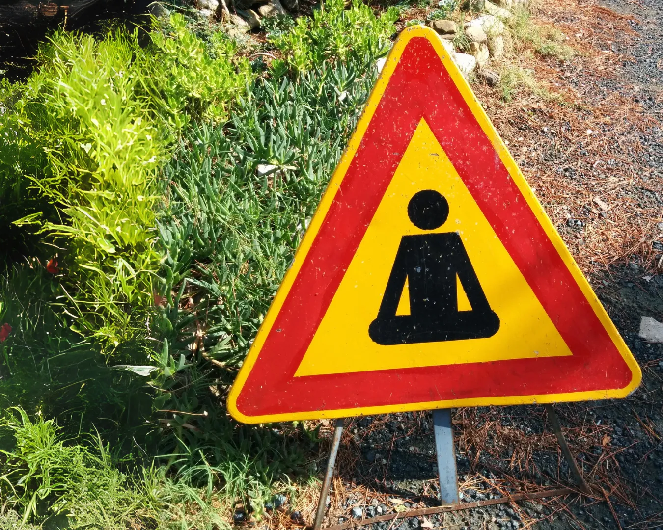 Cartello di attenzione triangolare con simbolo di meditazione yoga, posizionato lungo un sentiero naturale tra piante e ghiaia.