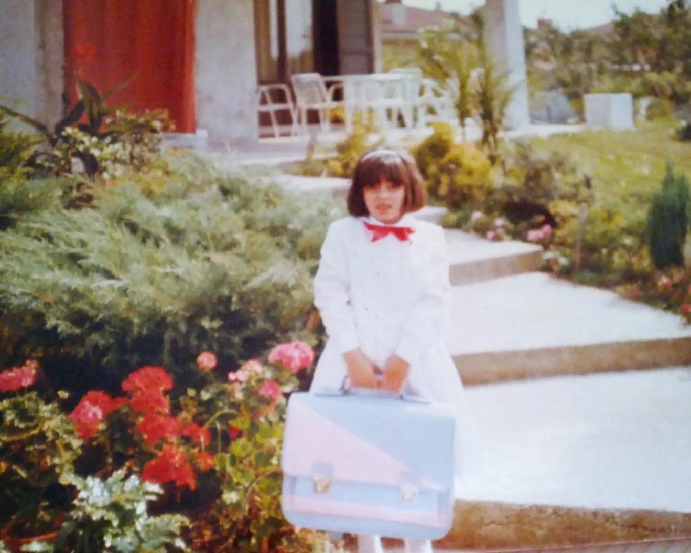 Chiara De Servi bambina nel suo primo giorno di scuola, con cartella in mano e vestita di bianco davanti a un giardino fiorito