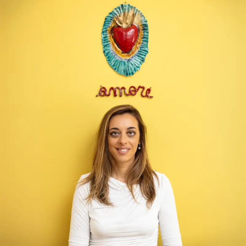 Claudia de Servi in piedi appoggiata a un muro giallo con la scritta amore sopra e una decorazione con cuore